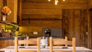 A rustic wooden interior with a dining table set for two, a hanging light fixture, and a kitchen area with wooden cabinets and a kettle. - Luxury Safari Lodge-Sleeps 4/6-Deer Park-Dogs Ok (Crediton)
