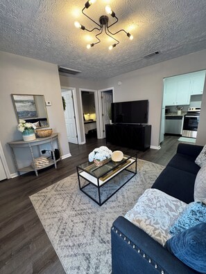 A living room with a sofa, coffee table, TV, and kitchen area. - Across from Campus! Magnolia LinenCo #7 Downtown AU (Auburn)