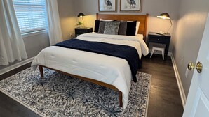 A bedroom with a bed, two bedside tables, a nightstand, a window with blinds, and a ceiling light fixture.