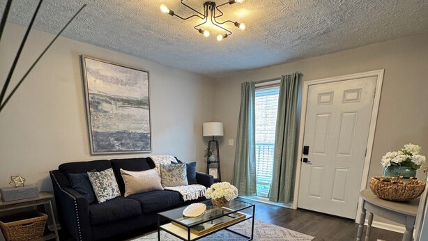 A living room with a black sofa, a glass coffee table, a patterned rug, a ceiling light fixture, and a door with a window.