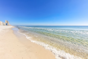 Ein Strand mit klarem Wasser, einem sandigen Ufer und Gebäuden im Hintergrund.