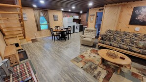 A cozy living area with a wooden floor, a sofa, a dining table with chairs, a ladder, and a kitchen in the background.