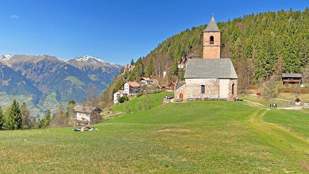 Une petite église en pierre dotée d’un clocher, entourée d’un champ verdoyant et de maisons, avec des montagnes en arrière-plan.