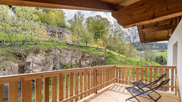 Une terrasse en bois dotée d’une rambarde, deux fauteuils de jardin noirs et une vue sur une colline parsemée d’arbres et bordée d’un mur de pierres.