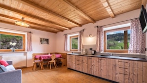 Une cuisine avec des meubles en bois, un coin repas avec une table et des chaises, et une vue sur un paysage verdoyant grâce à de grandes fenêtres.