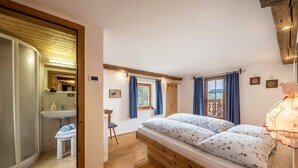 Ein Schlafzimmer mit Bett, Nachttisch, Stuhl, einem Fenster mit Vorhängen, Holzboden und Blick ins Freie.