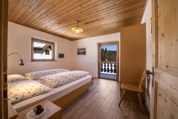 A bedroom with a wooden ceiling, a bed with floral bedding, a small table with a decorative item, a window with a view, and a door leading to a balcony.