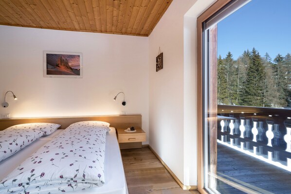 A bedroom with two beds, a wooden ceiling, a nightstand, a wall-mounted picture, and a balcony with a railing.