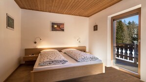Ein Schlafzimmer mit Holzbalkendecke, einem Bett mit floralem Bettzeug, einem Nachttisch, einer Wandleuchte und einem Balkon mit Geländer.