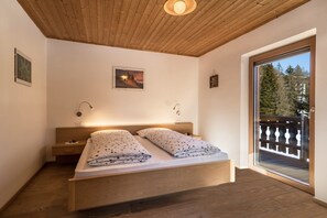 A bedroom with a wooden ceiling, a bed with floral bedding, a nightstand, a wall-mounted lamp, and a balcony with a railing.