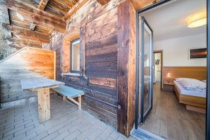 Interior de una cabaña de madera con mesa, banco y cama con cabecera.