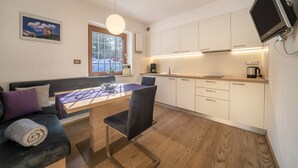 A modern kitchen with white cabinets, a dining table with chairs, a built-in TV, and a window with a view of mountains. - Apartment "Hirzer" near Skiing Area with Forest View, Wi-Fi, Balcony & Shared Garden (Falzeben)