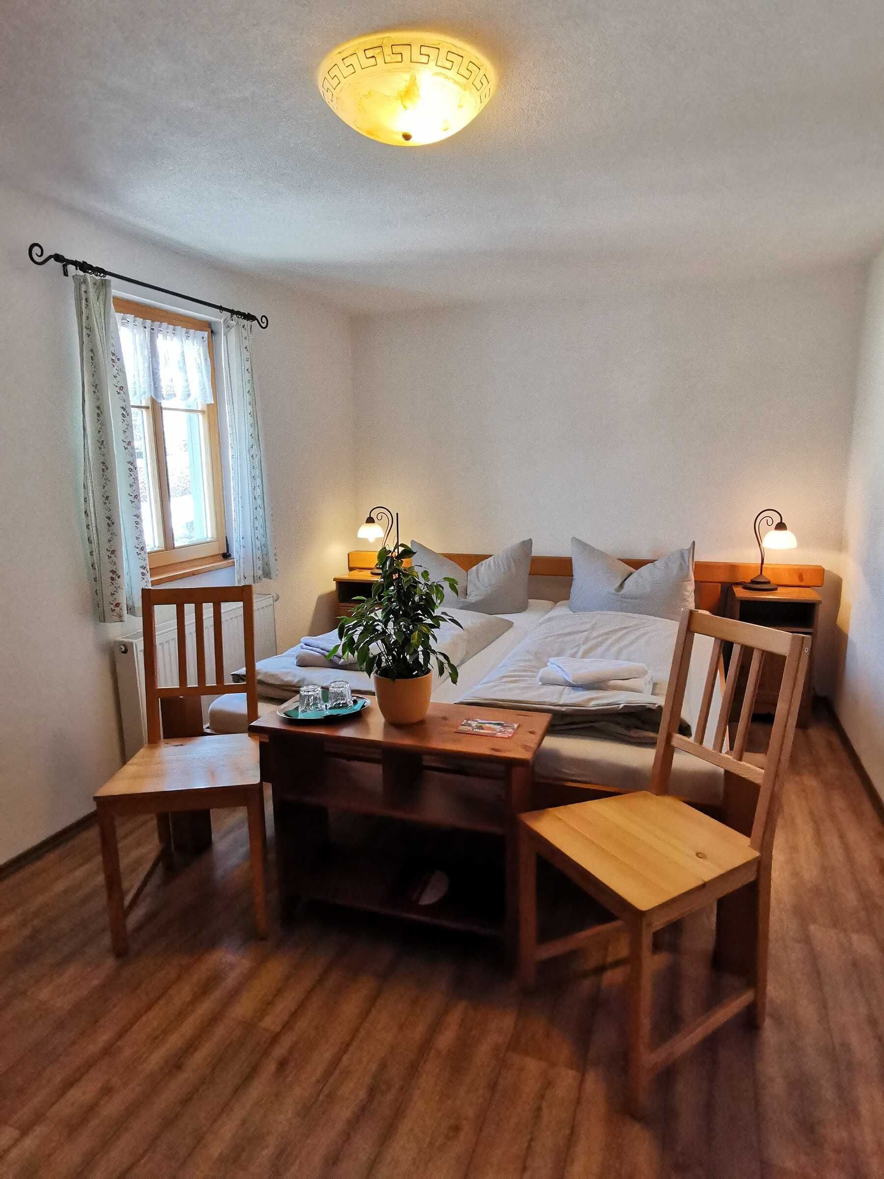 Een gezellige kamer met een bed, twee nachtkastjes met lampen, een houten stoel, een kleine tafel met een plant en een raam met gordijnen.