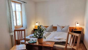Ein gemütliches Zimmer mit einem Bett, zwei Nachttischen mit Lampen, einem Holzstuhl, einem kleinen Tisch mit Pflanze und einem Fenster mit Vorhängen.