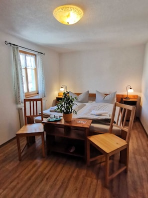 Un cuarto acogedor con una cama, dos mesitas de noche con lámparas, una silla de madera, una pequeña mesa con una planta y una ventana con cortinas.
