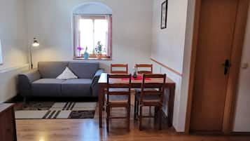 Ein gemütliches Wohnzimmer mit grauem Sofa, einem Holzesstisch und Stühlen. Ein Fenster mit Vorhängen, ein Heizkörper und eine Tür sind ebenfalls zu sehen.