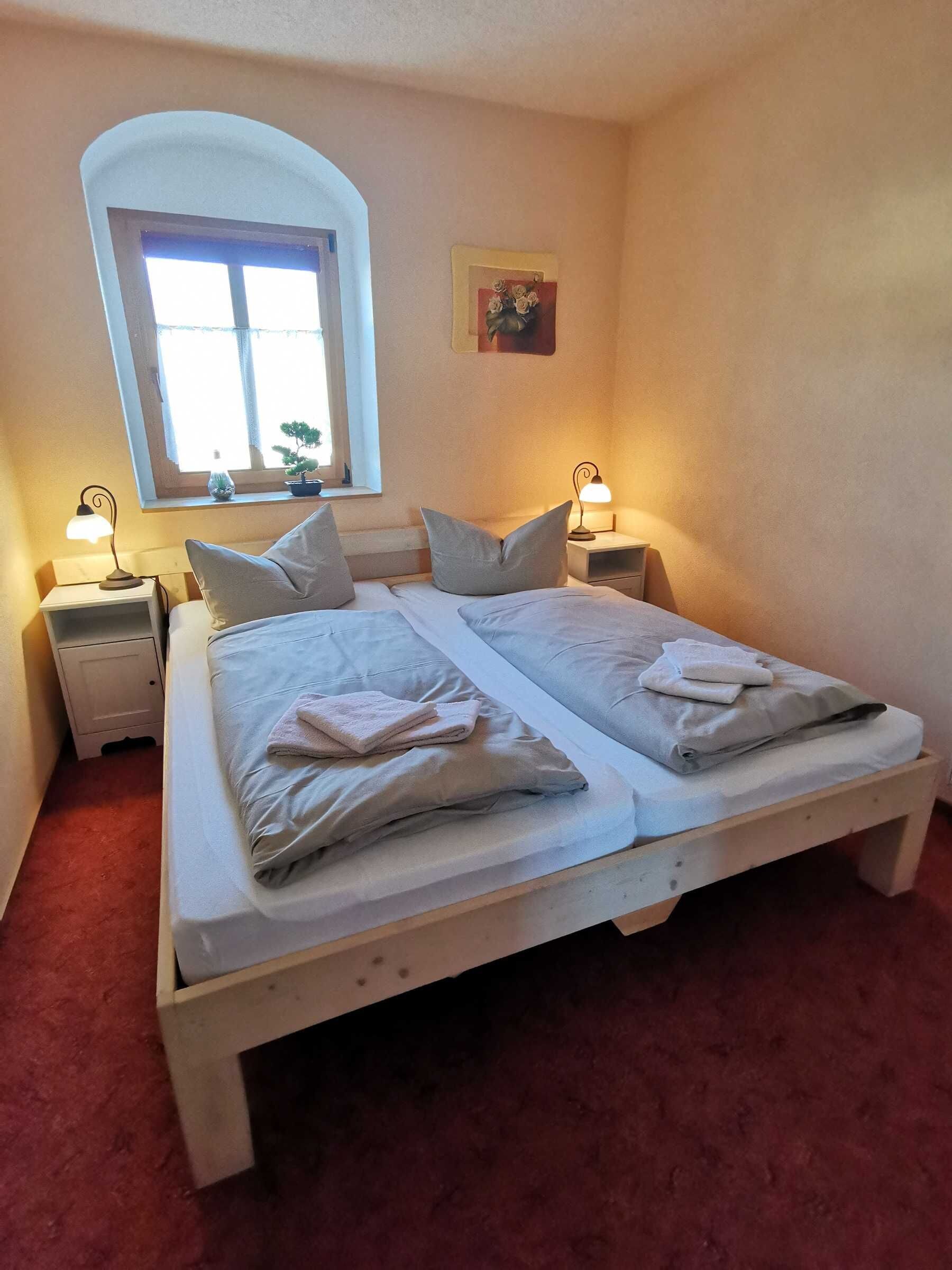Ein Doppelbett mit zwei Kissen, ein Nachttisch mit Lampe, ein Fenster mit Blick auf Grünflächen und ein kleines Bild an der Wand.