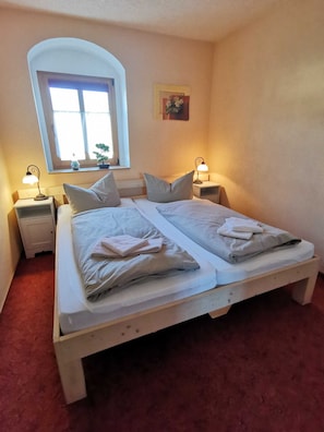 Una cama doble con dos almohadas, una mesita de noche con lámpara, una ventana con vistas al verdor y un cuadro pequeño en la pared.