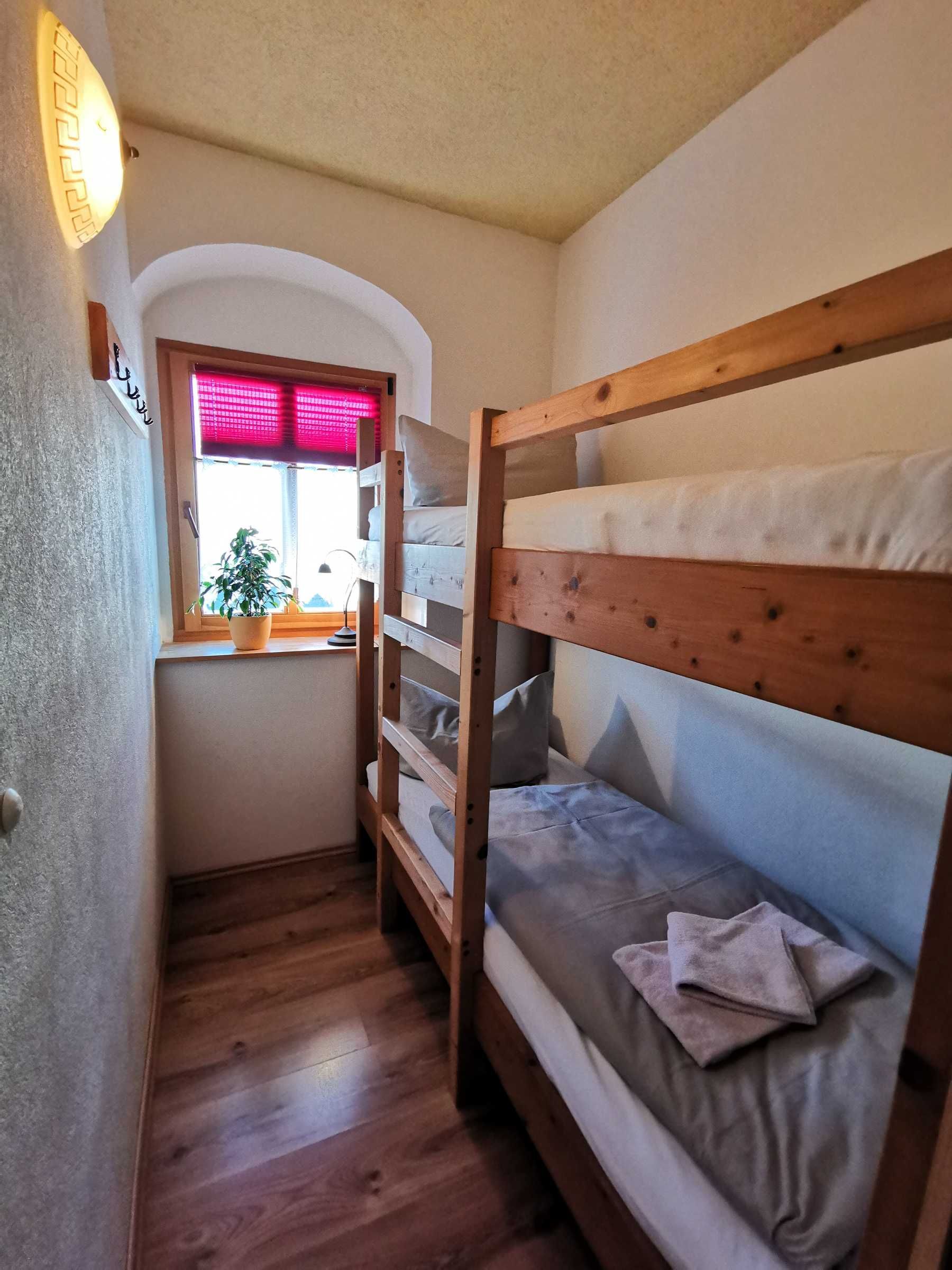 Ein kleiner, gemütlicher Raum mit einem Etagenbett, einem Fenster mit rotem Rollo und einem Blumentopf am Fenster.