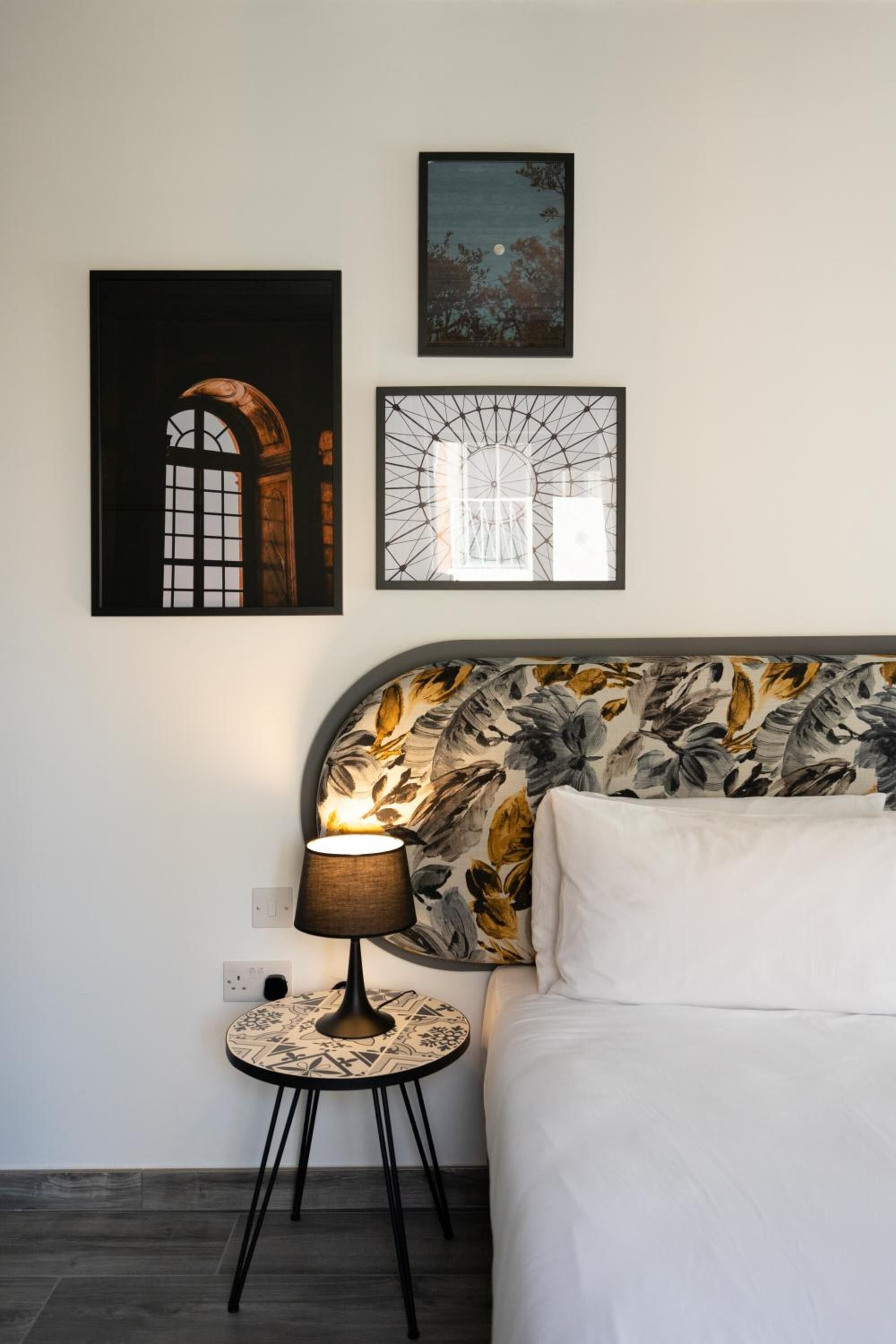 Een slaapkamer met een bed, een ronde tafel met een lamp en twee kunstwerken in lijsten aan de muur.