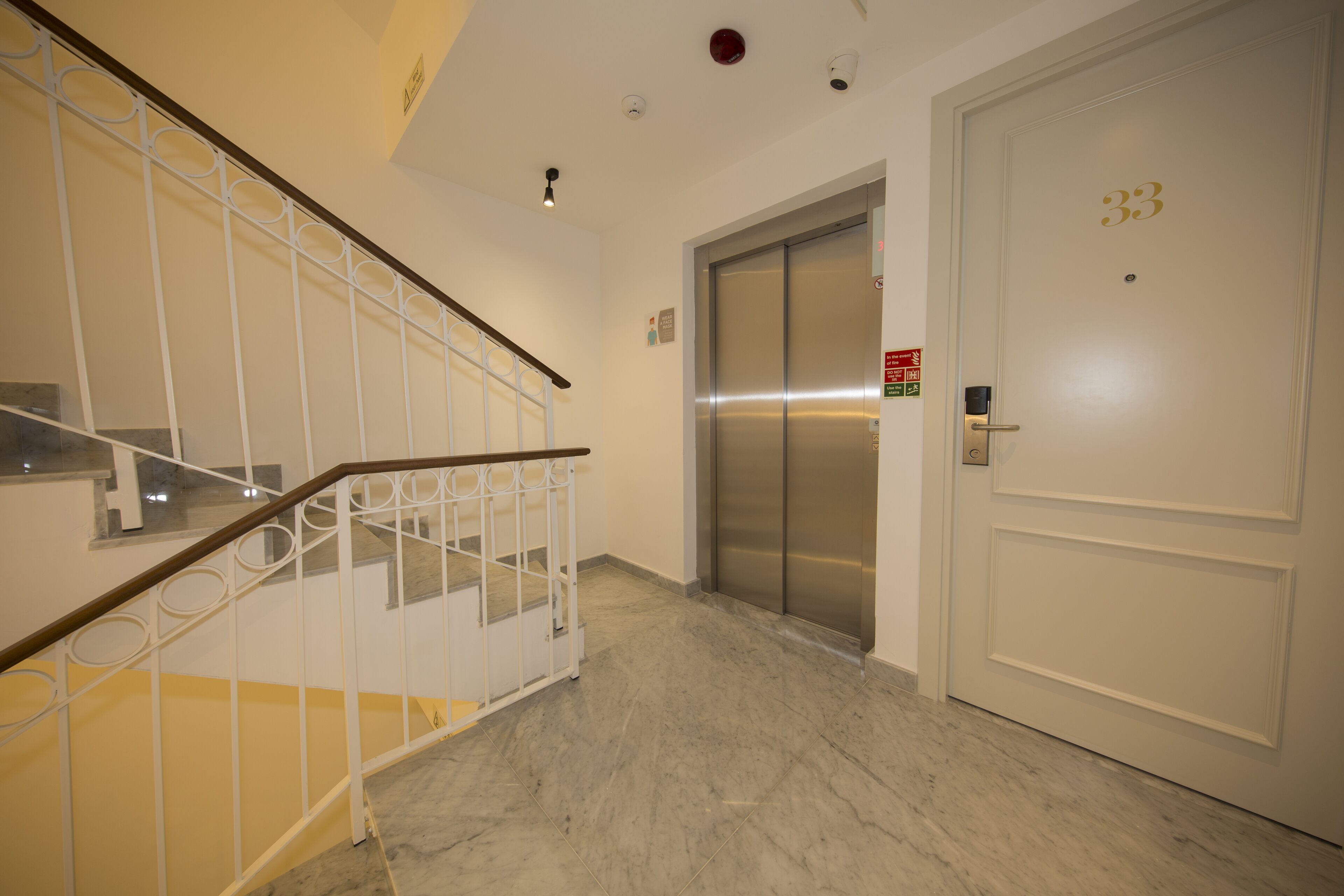 Un ascensor moderno y limpio con detalles metálicos, una puerta blanca con el número 33, y una escalera con barandilla blanca y escalones amarillos.