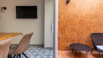 Una habitación con una mesa de madera, dos sillas acolchadas, un televisor de pantalla plana en la pared y una mesita con una silla.