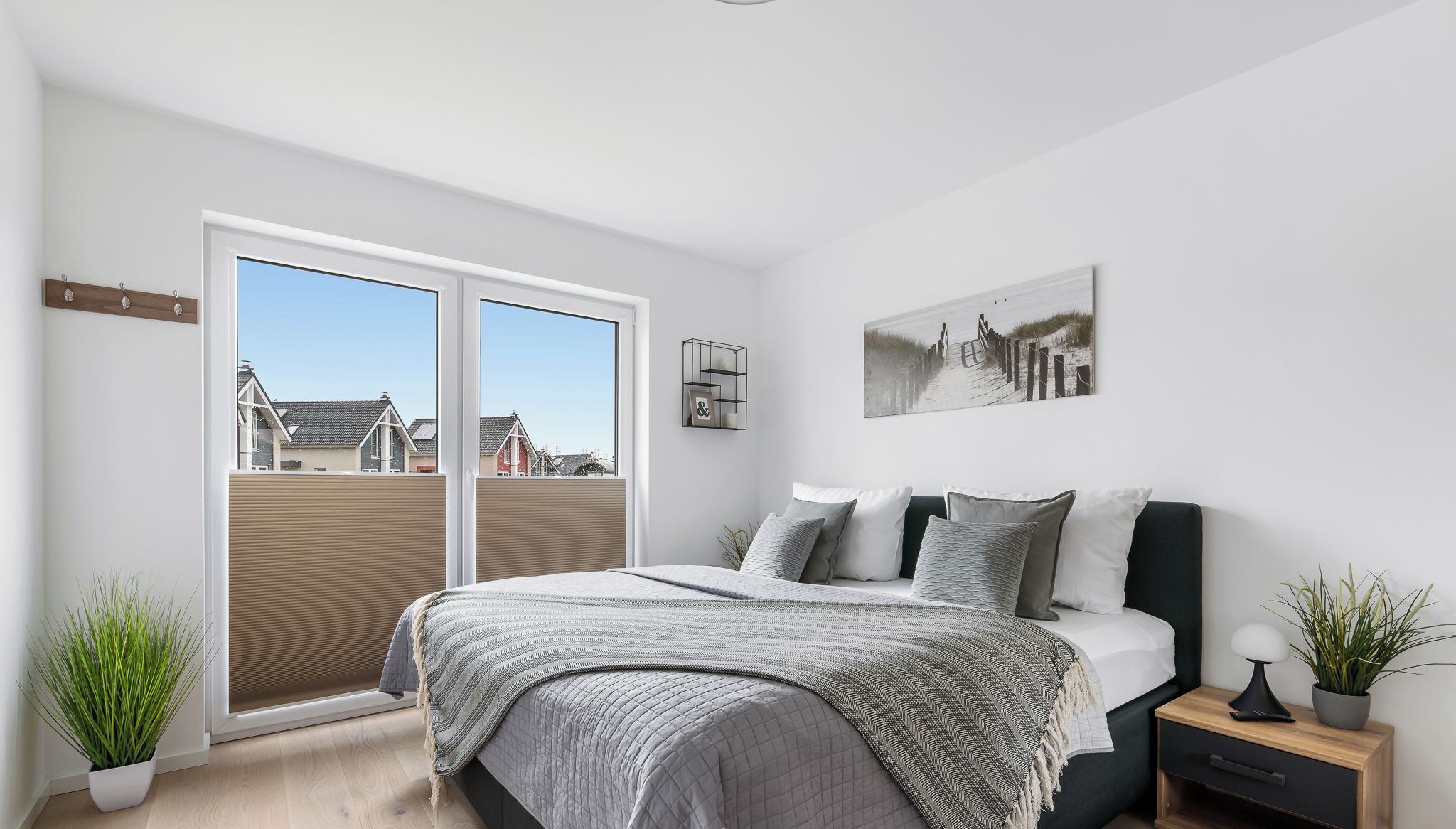 Un dormitorio con cama, mesita de noche, planta en maceta y ventana con vistas a casas.