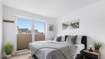 Un dormitorio con cama, mesita de noche, planta en maceta y ventana con vistas a casas.
