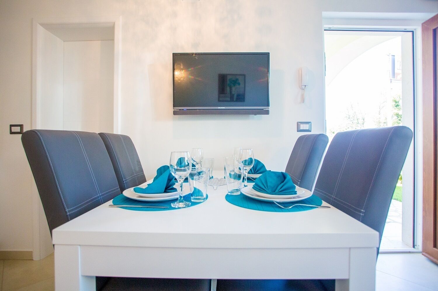 Un coin repas avec une table blanche, deux chaises bleues à revêtement rembourré et un téléviseur à écran plat fixé au mur.