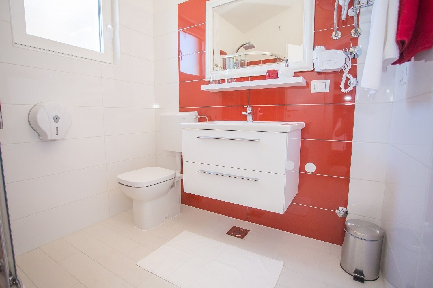 A bathroom with a white toilet, a white sink with a red-tiled wall, a mirror above the sink, and a trash bin to the right.