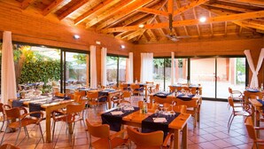 Un comedor amplio con vigas de madera, ventanas grandes y sillas color naranja. Las mesas están preparadas con manteles blancos y cristalería.