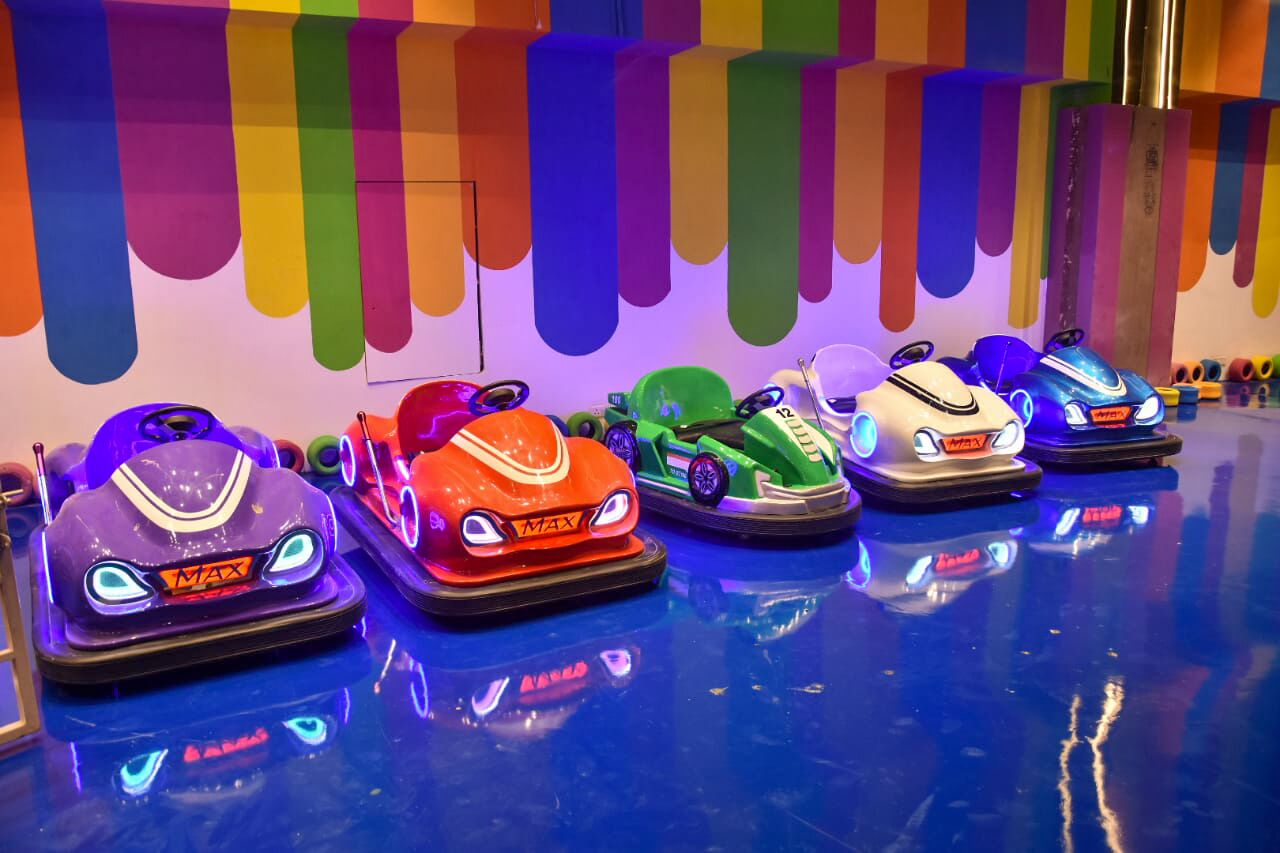 A row of colorful bumper cars with the word "MAX" on them, parked on a reflective floor with a colorful wall in the background.