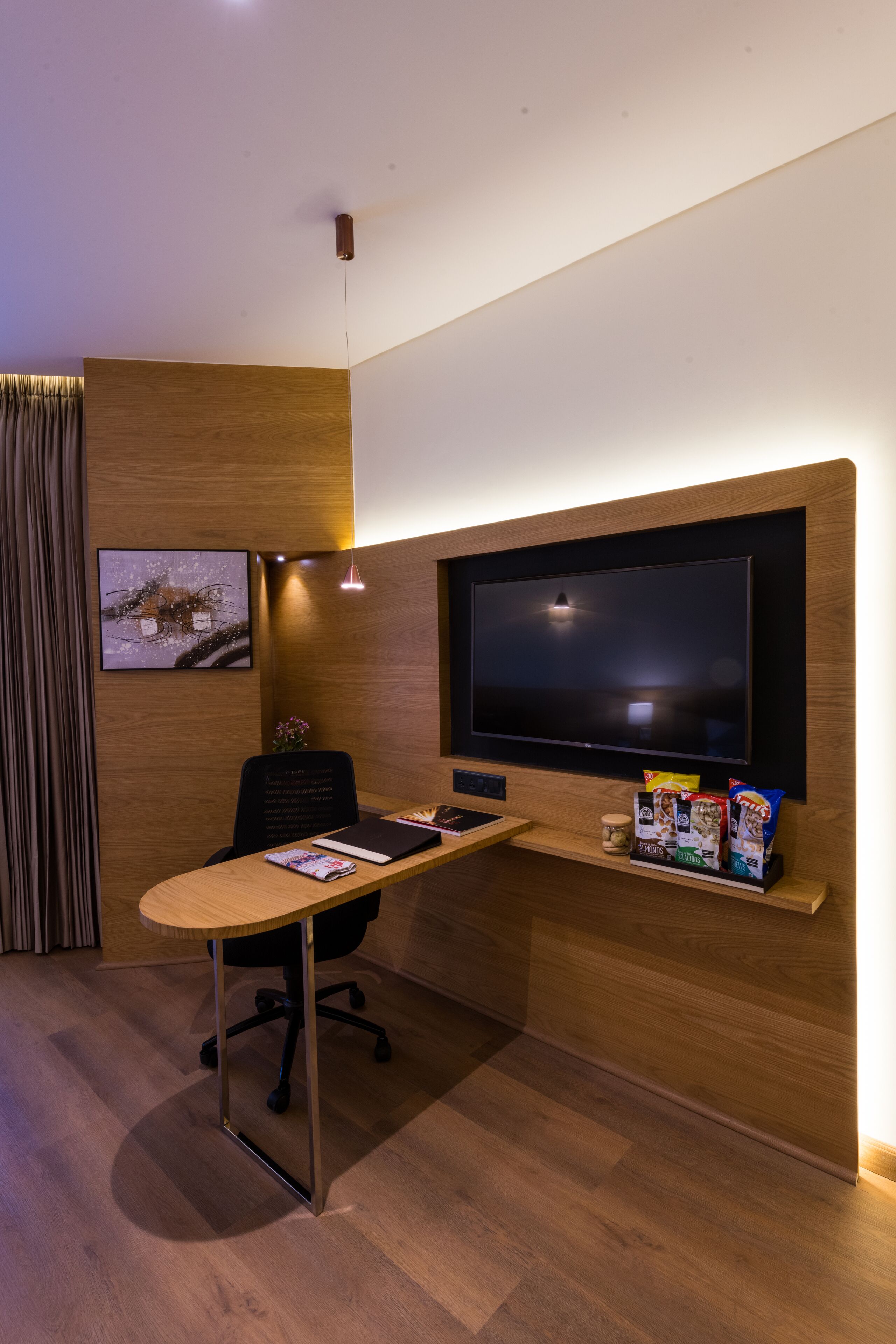 A modern office space with a large flat-screen TV mounted on a wooden wall, a desk with a chair, and a shelf with snack packages.