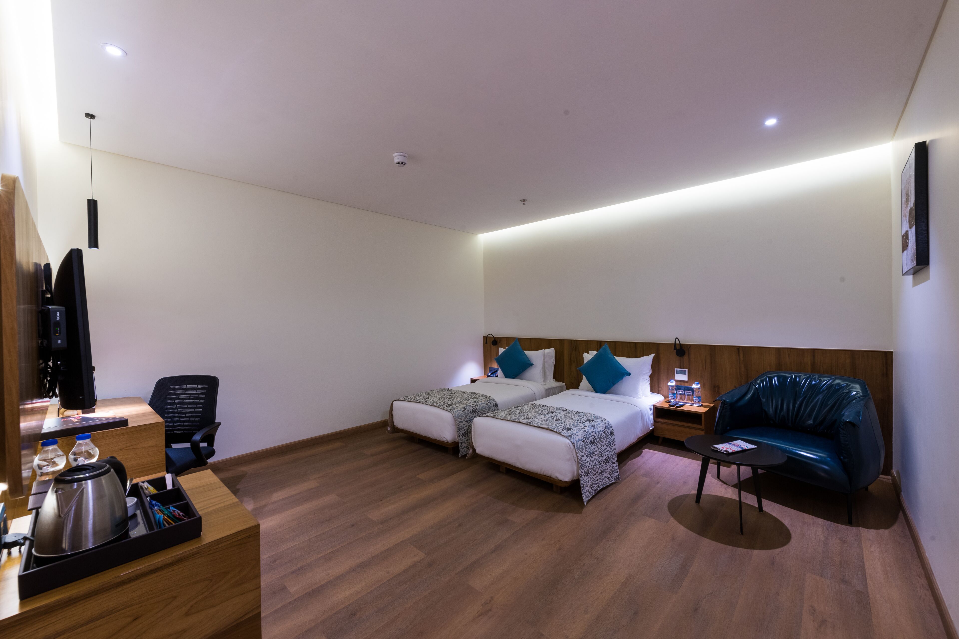 A hotel room with two beds, a desk with a kettle, a chair, a small table, and a blue sofa.