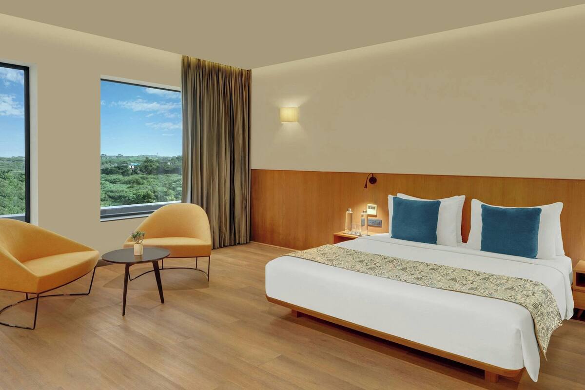A hotel room with a large bed, two armchairs, a small table, and a view of greenery through the window.