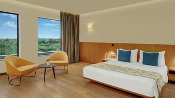 A hotel room with a large bed, two armchairs, a small table, and a view of greenery through the window.