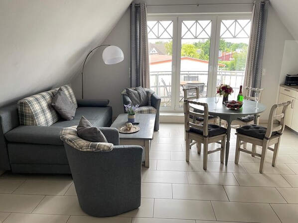 Una sala de estar moderna con sofá, sillón, mesa de comedor y sillas. Se observa una ventana grande con cortinas, y una lámpara es visible.