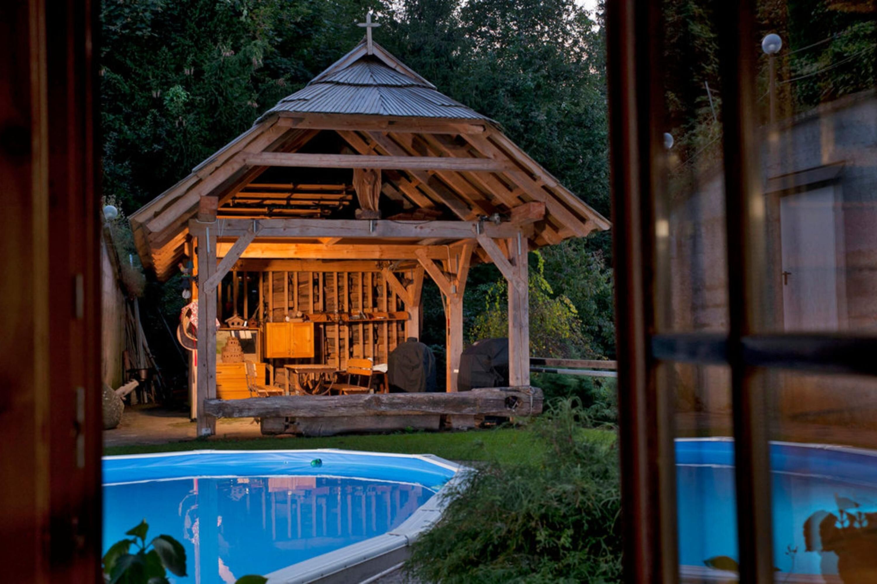 Ein hölzernes Pavillon mit Dach, umgeben von viel Grün, und ein Schwimmbecken im Vordergrund.