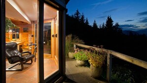 A modern house with a glass door leading to a living area, a wooden deck with potted plants, and a view of trees and mountains.
