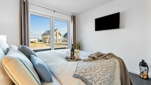 A bedroom with a large window, a bed with pillows, a bedside table with a bottle and glasses, and a TV on the wall.