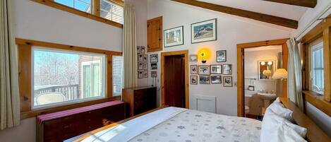 A bedroom with a bed, wooden beams, a window, a bench, and a wall with framed pictures.