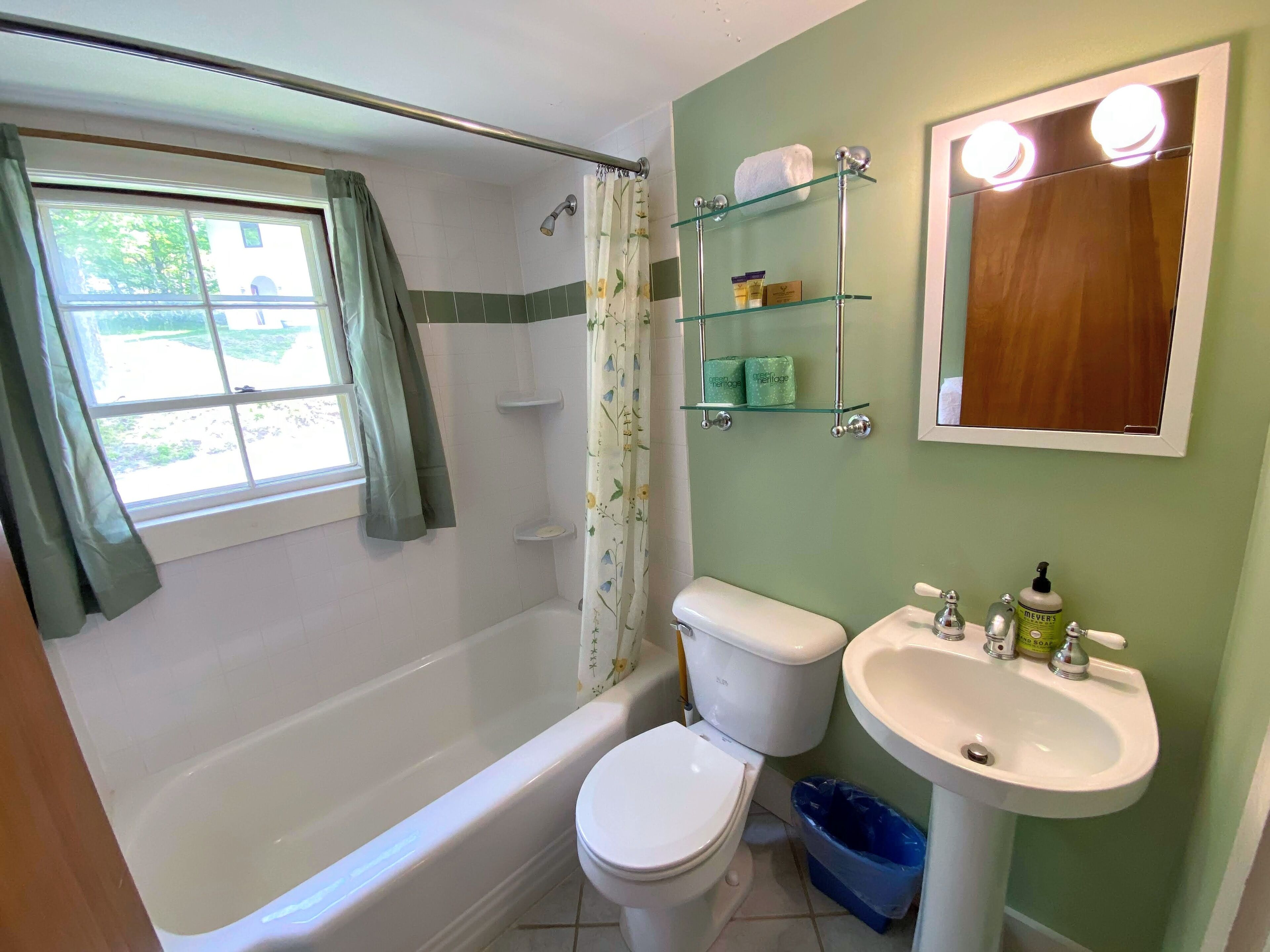 A bathroom with a bathtub, toilet, and sink. There is a window with curtains, a shower curtain, and shelves with toiletries.