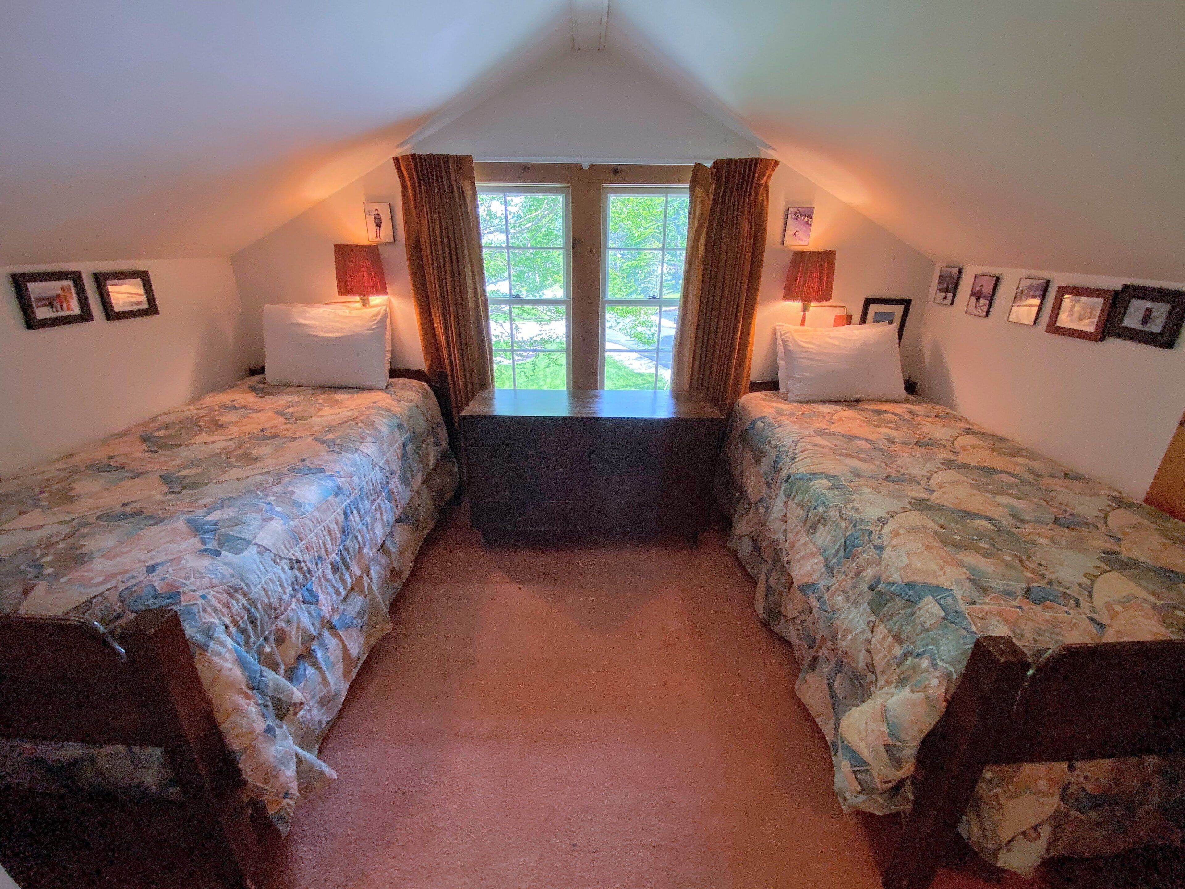 A room with two beds, a window with curtains, and framed pictures on the walls.