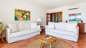 Uma sala de estar com dois sofás brancos, uma mesa de centro de madeira e um quadro abstrato colorido.
