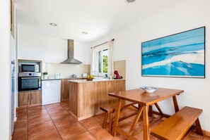 A compact kitchen with a central island, a dining table with benches, and a large abstract painting on the wall. - Villa Casa Nova, Piscina, Wifi, AC gratis (Sant Lluís)