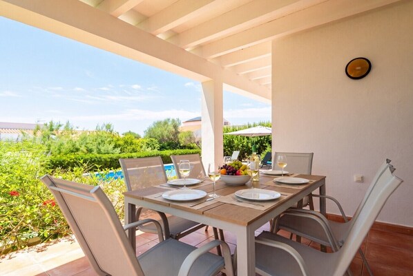 An outdoor dining area with a table set for four, surrounded by chairs, with a view of a pool and greenery. - Villa Casa Nova, Piscina, Wifi, AC gratis (Sant Lluís)