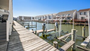 A waterfront property with a wooden dock extending into a calm waterway, surrounded by residential buildings with balconies and decks. - Sea Robin 207 by Seaside Vacations & Sales (Ocean City)