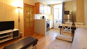 Ein kompakter Wohnraum mit Fernseher, einer Bank, einem Sofa, einer Küchenzeile inklusive Waschmaschine sowie einem Essbereich mit Stühlen.