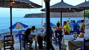 An outdoor dining area with tables and chairs, some covered by umbrellas, overlooking a body of water with cliffs in the background. - Beach Front Apartments (Sitia)