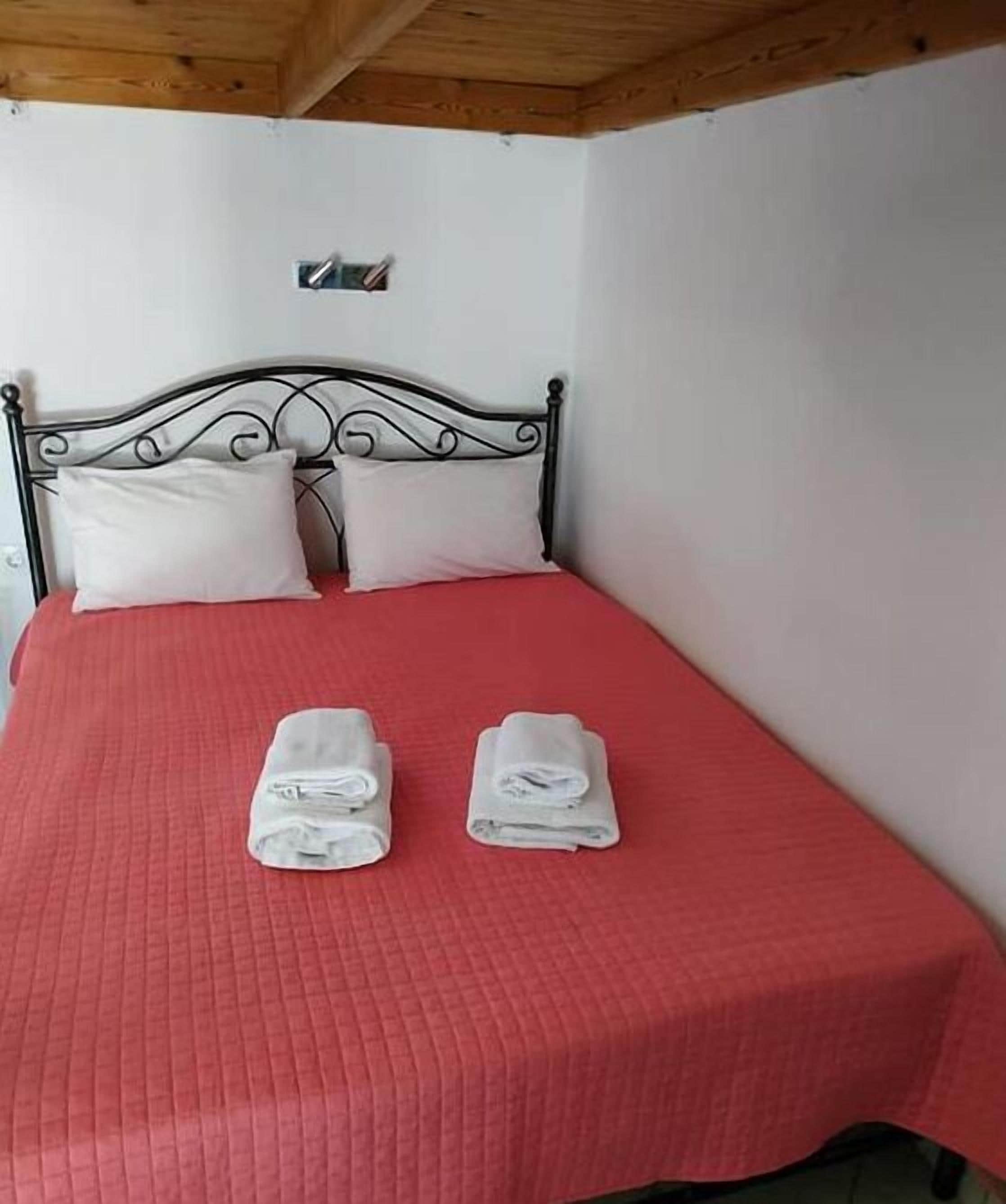 A single bed with a red bedspread, two pillows, and two rolled towels on the bed.